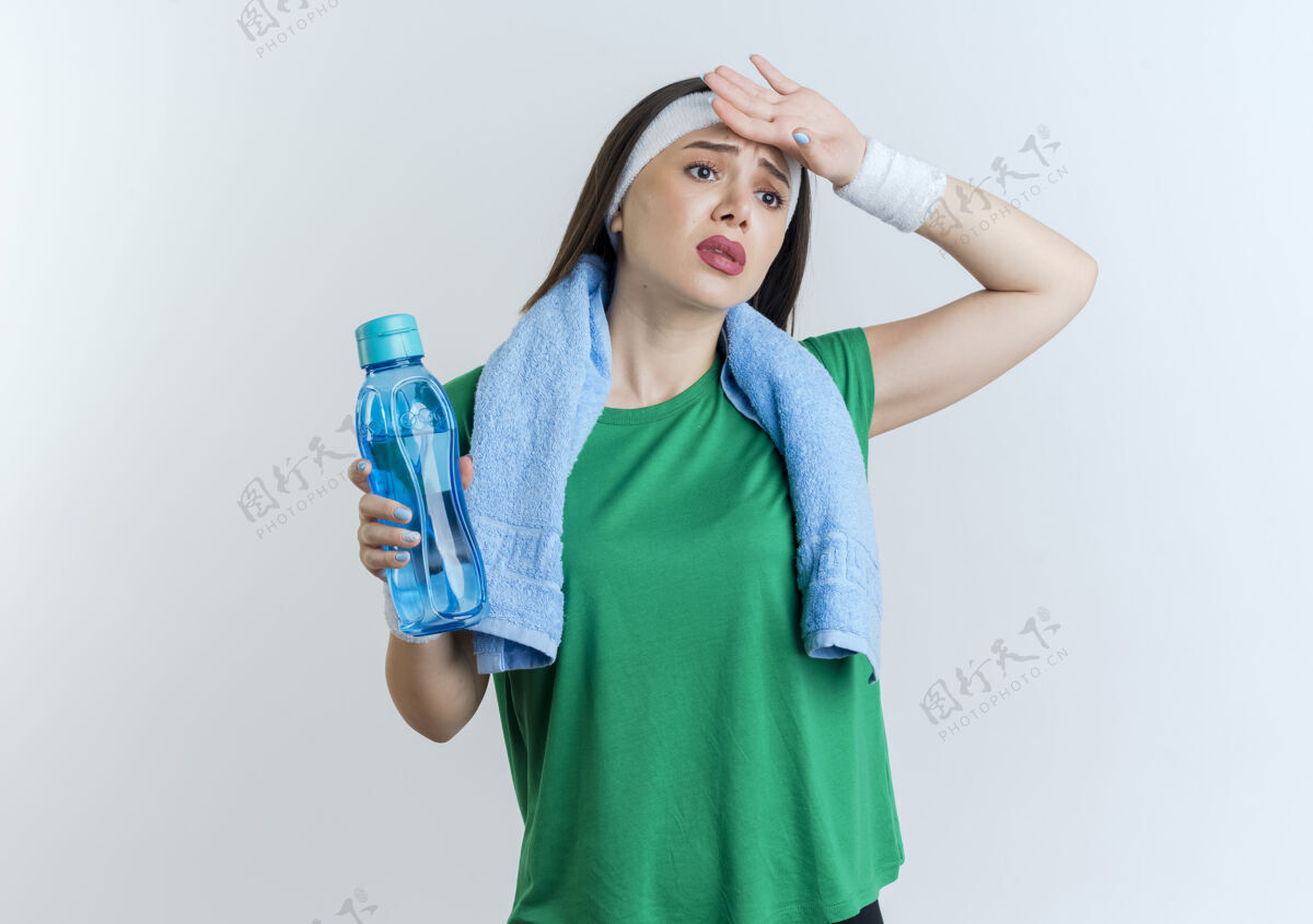 运动疲惫的年轻运动女性戴着头带和腕带 脖子上围着毛巾 拿着水瓶 手放在额头上 看着侧面运动放脖子