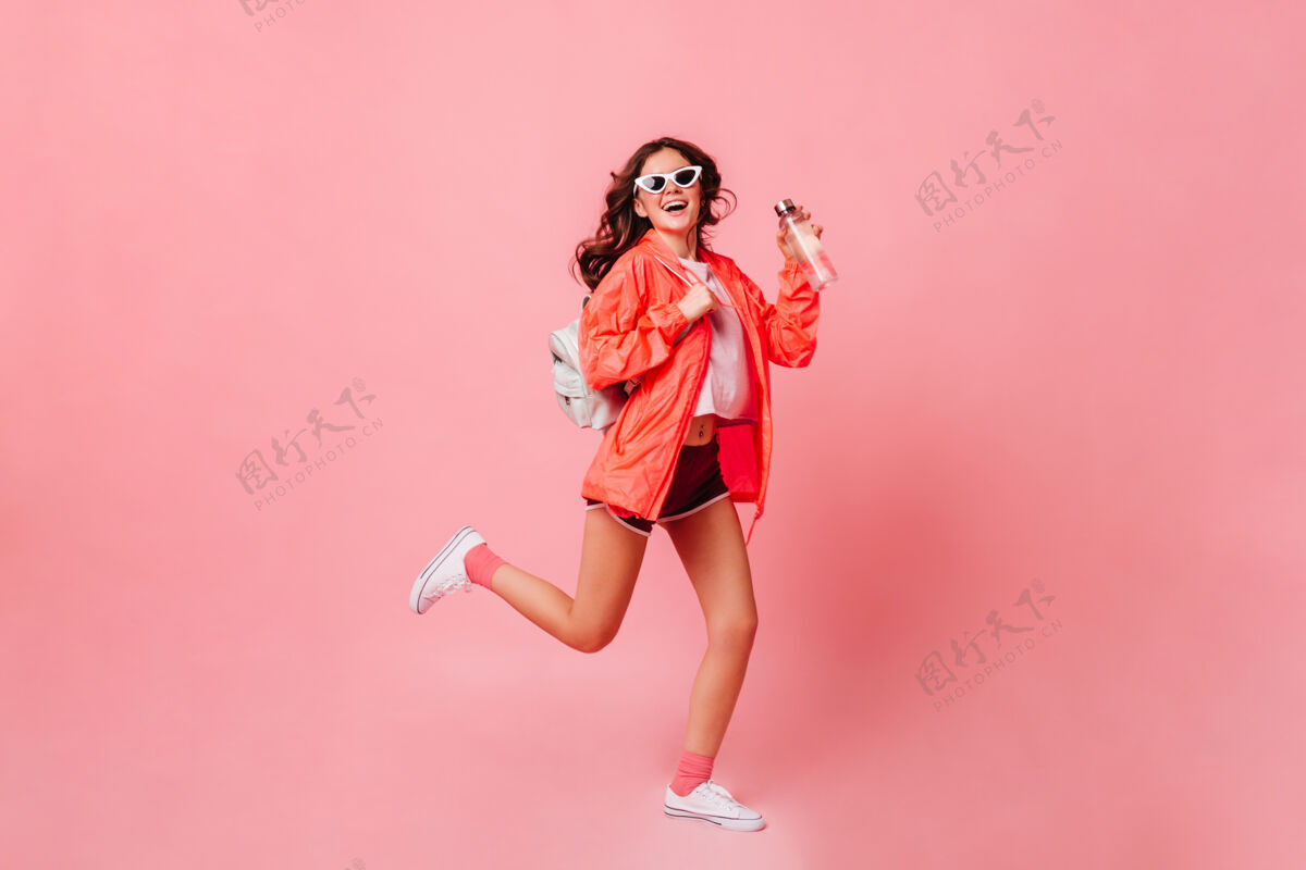 漂亮穿着风衣 短裤和运动鞋的女运动员在粉红色的墙上跑步脸太阳镜女士