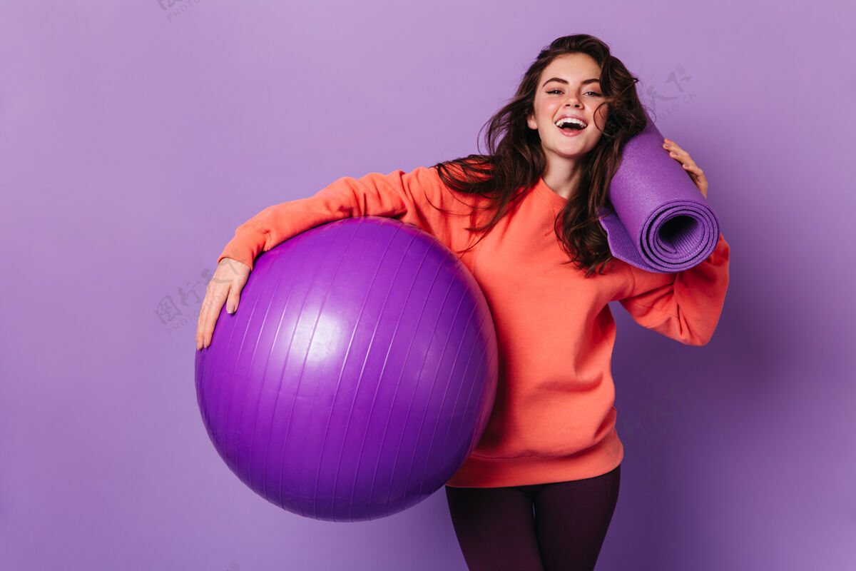 人穿着紧身裤和亮色运动衫的美女微笑着摆出紫色垫子和健身球的姿势成人肖像锻炼