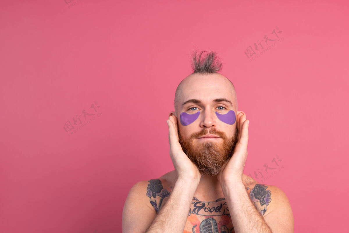 脸欧洲帅哥留着胡子纹身上身的男人戴着紫色眼罩对着镜头摆粉红色的姿势清洁清洁治疗