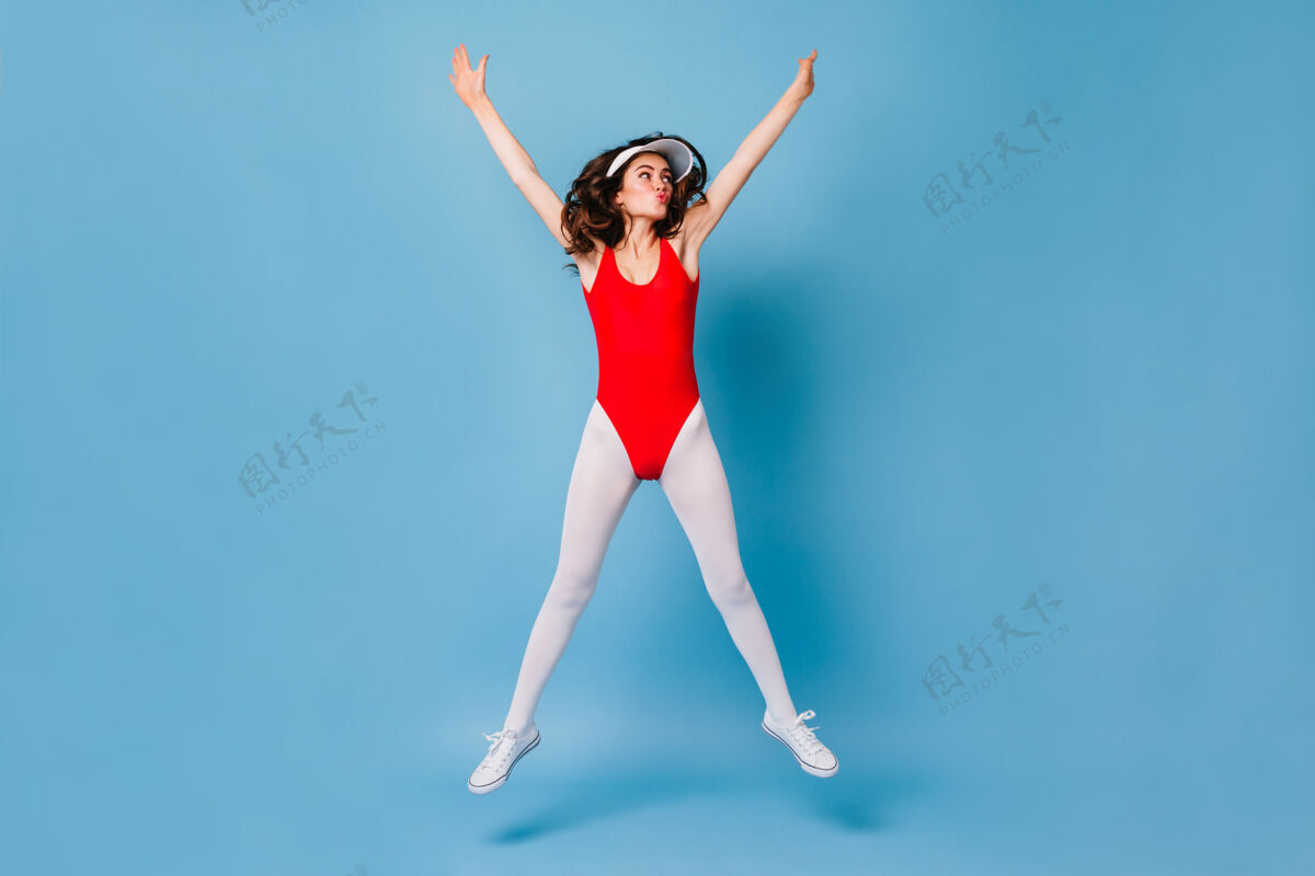 80后风格身穿红色运动紧身衣和白色紧身裤的苗条黑发女子在蓝色的墙上跳跃皮肤时尚美容
