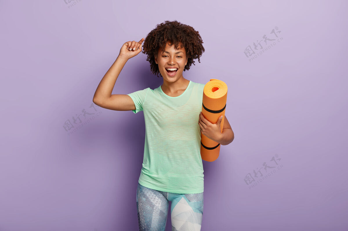 穿着喜出望外的女人拿着瑜伽垫 举起手臂 穿着休闲t恤和紧身裤 对积极的锻炼感到高兴女性锻炼卷发