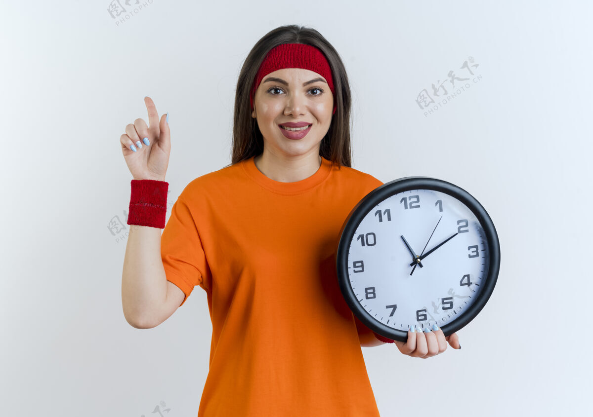 腕带高兴的年轻运动女子戴着头带和腕带拿着时钟抬头看运动年轻向上