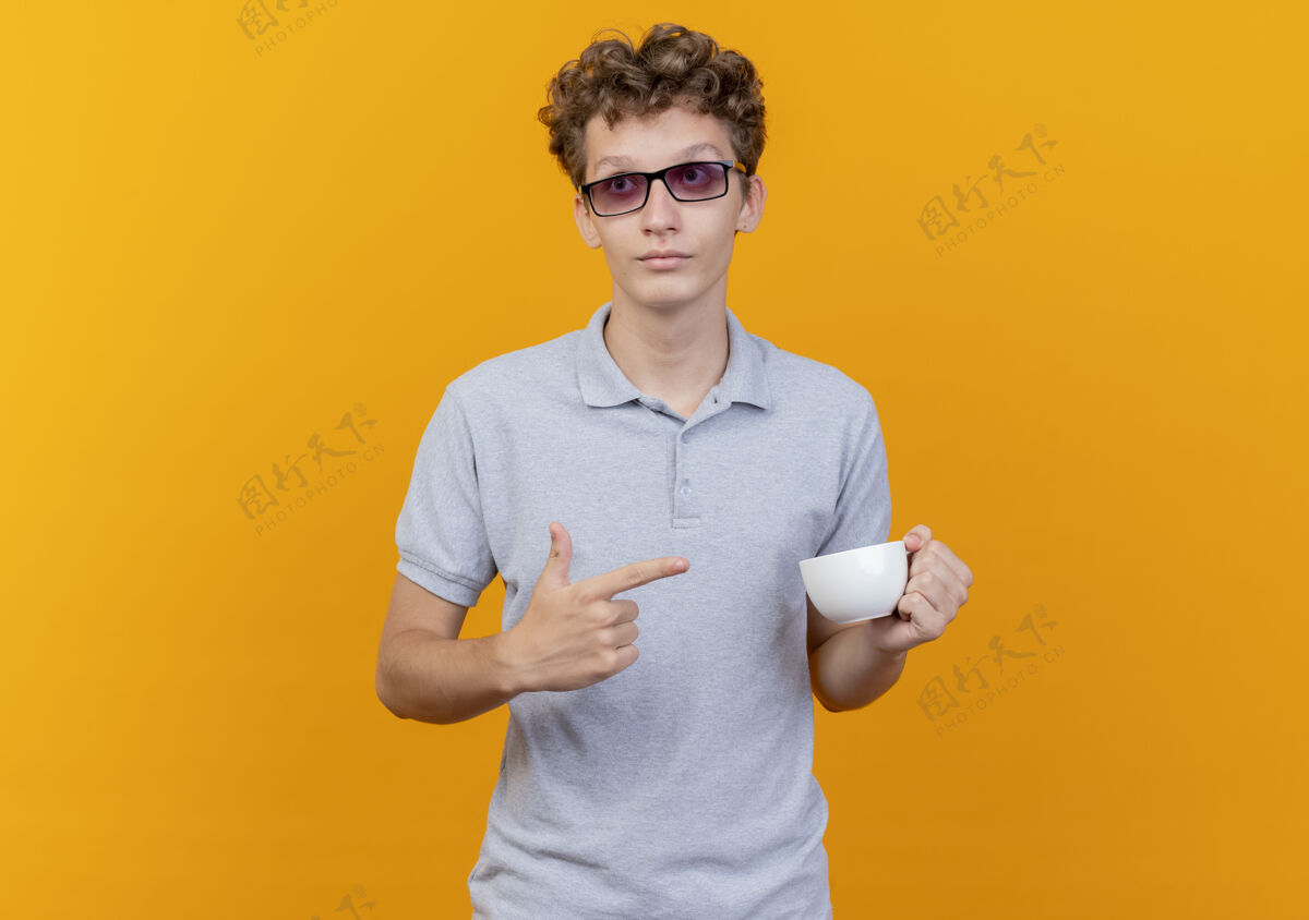脸戴着黑眼镜的年轻人 穿着灰色马球衫 手里拿着咖啡杯 用手指着咖啡杯 严肃地看着绿色的脸看靠边黑色