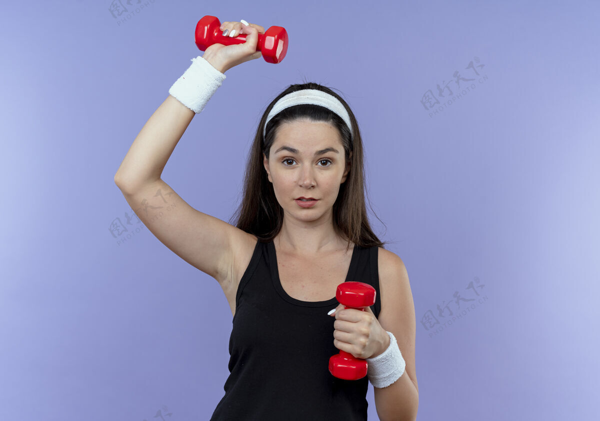 工作戴着头巾的年轻健身女士站在蓝色的墙上 带着哑铃 看起来很自信长相年轻健身
