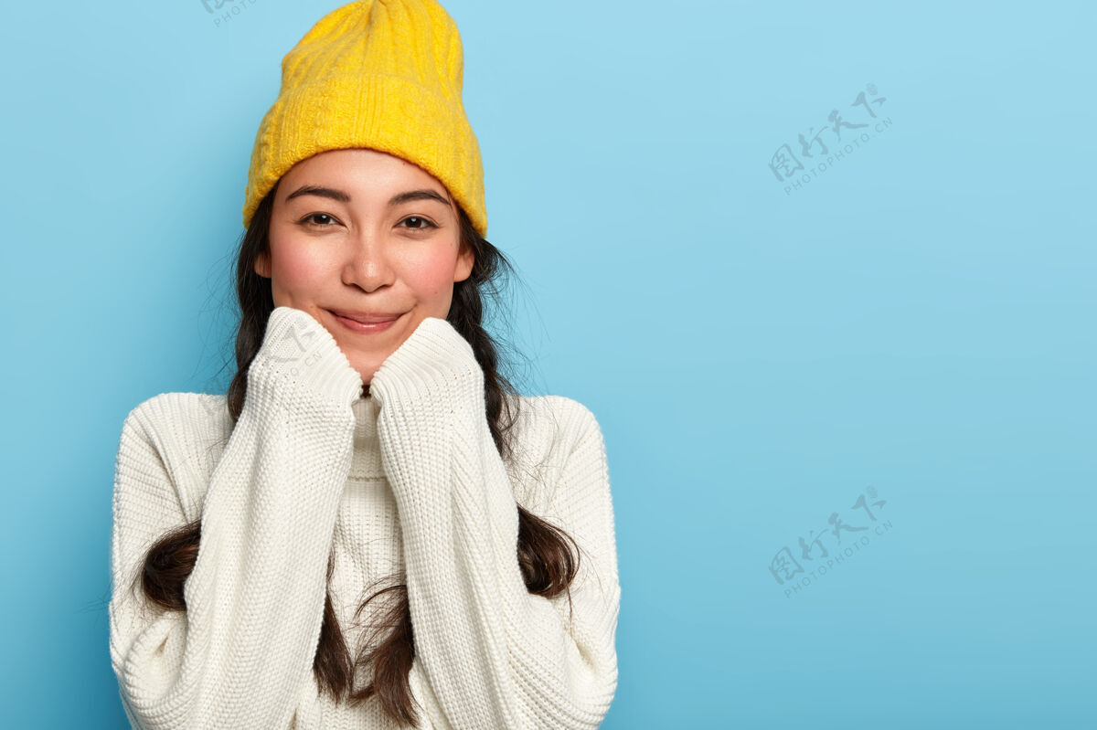 肖像快乐可爱的亚洲女人的画像 手放在下巴下 穿着超大的白色毛衣和黄色帽子 享受舒适和平静的家庭气氛温暖微笑民族