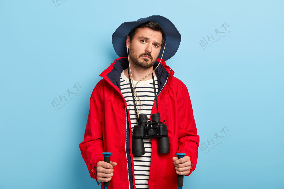 白天严肃的年轻男性徒步旅行者在树林里旅行 使用登山杖 享受旅游生活方式 脖子上戴着望远镜 戴着休闲帽和红色夹克 隔离在蓝色的墙上旅行站立徒步旅行