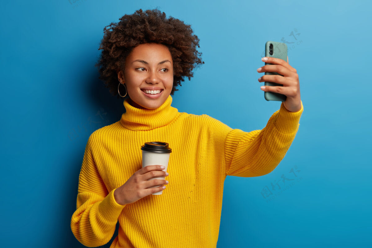 高兴快乐的非洲女孩在网上录制视频 在手机上自拍 用现代小玩意儿伸胳膊 拍照 拿着纸杯和咖啡视频通话美女女士