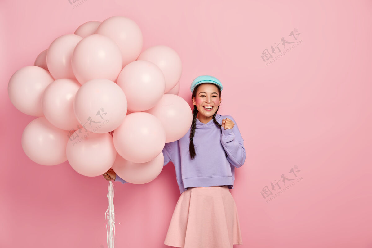 美女快乐的亚洲女孩高兴地握紧拳头 迫不及待地等待着特殊的时刻 在她的生日得到朋友们的祝贺 带着一大串气球 穿着漂亮的衣服模特编织女人