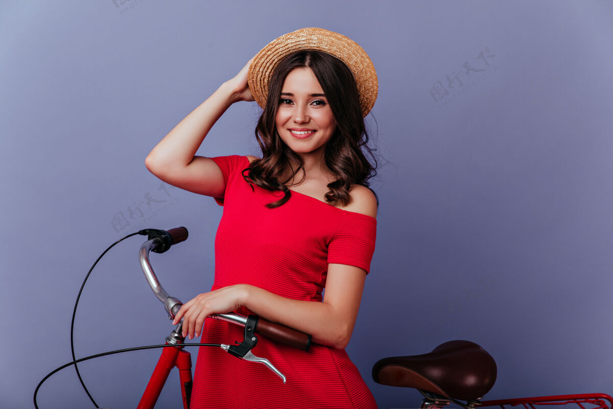 孤立的紫罗兰色戴着夏日帽子的迷人活泼的女孩坐在自行车上令人惊叹的白色女人在紫色内饰的自行车上摆姿势休闲活动活动自行车
