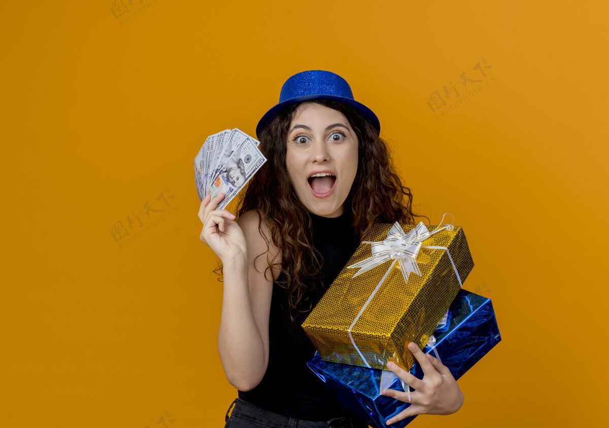 站着一个年轻漂亮的女人 卷发 戴着派对帽 手里拿着现金和礼物 看着卡内拉站在橘色的墙上 快乐而兴奋兴奋现金女人