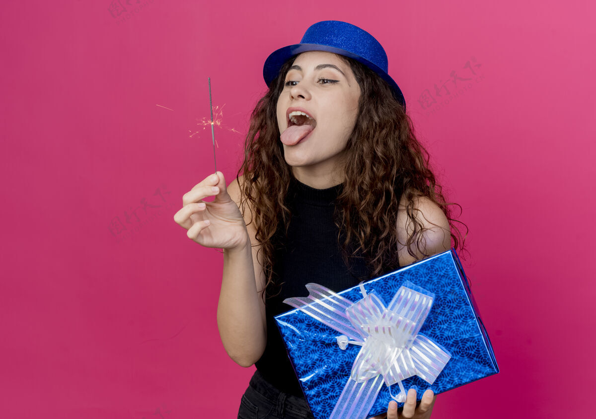 帽子年轻漂亮的女人 卷发 戴着节日礼帽 手里拿着生日礼盒 手里拿着闪闪发光的生日快乐派对概念站在粉色的墙上抱着闪闪发光疯狂