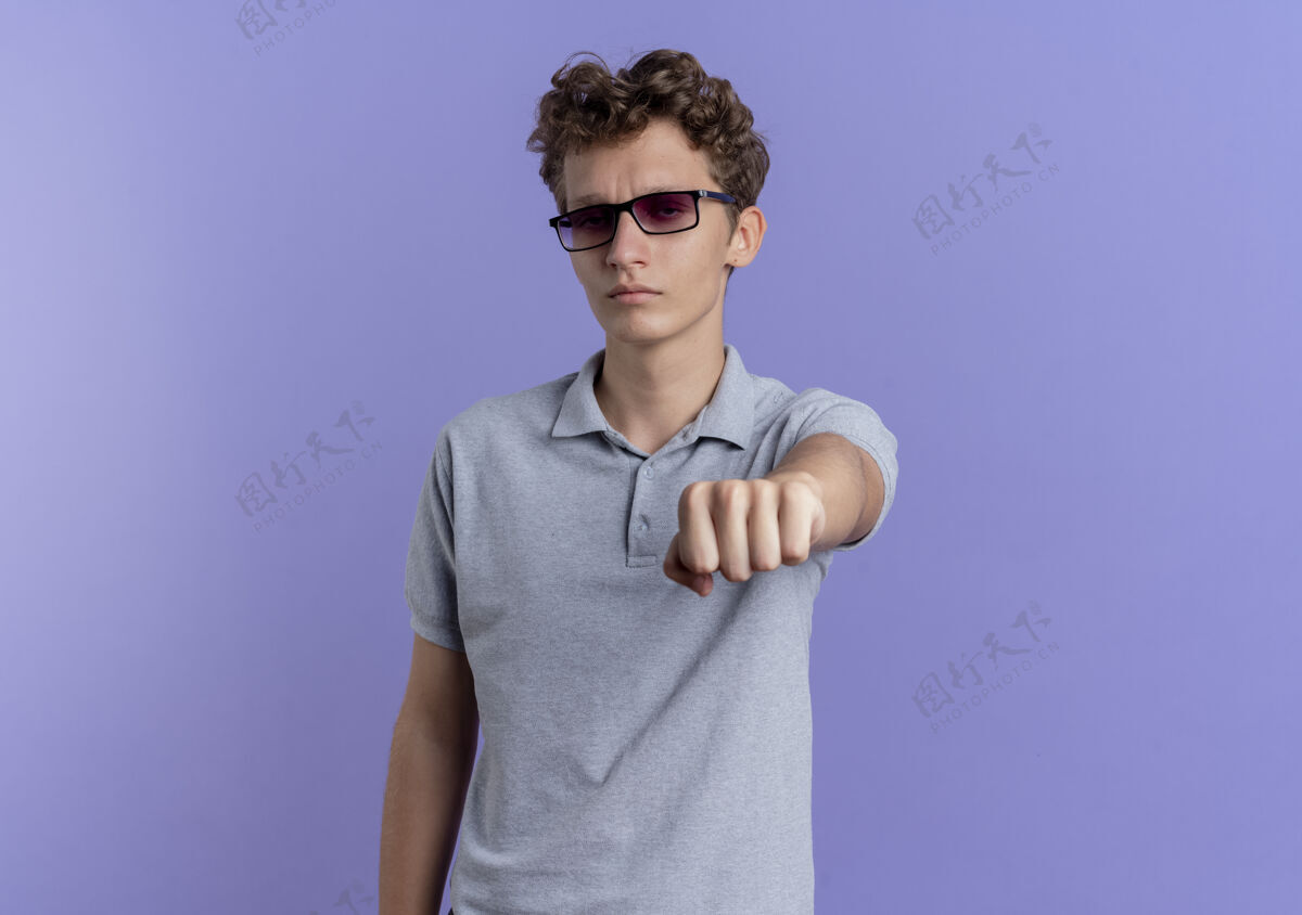 站着一个戴着黑眼镜 穿着灰色马球衫的年轻人站在蓝色的墙上 用拳头指着脸 表情严肃男人指着穿