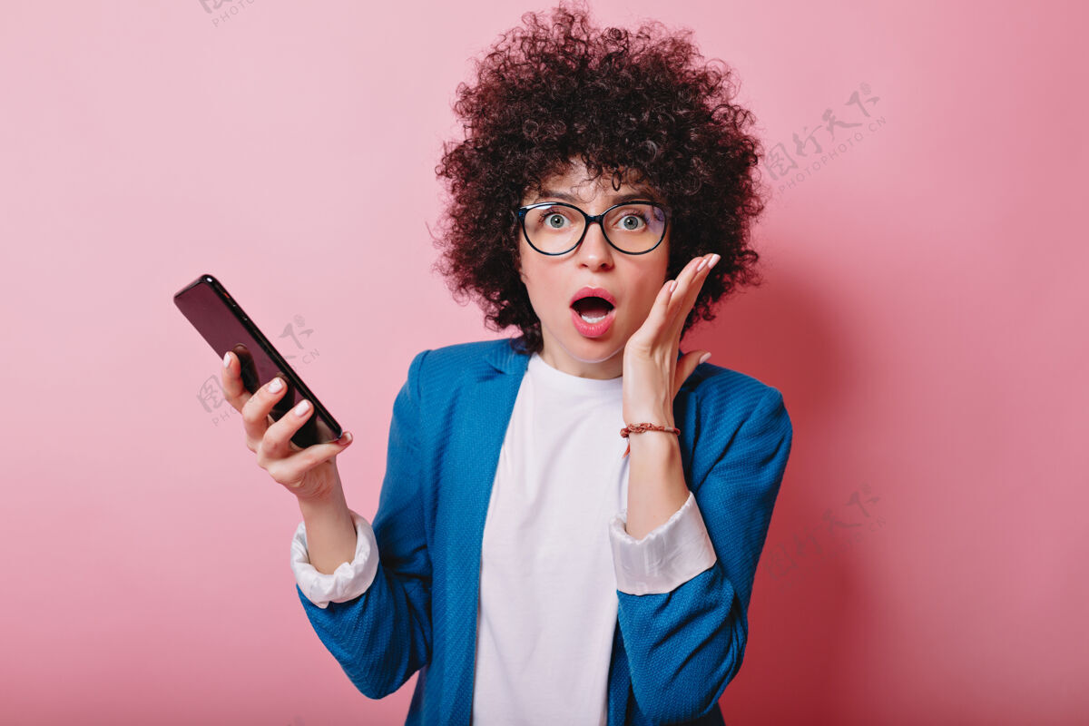 酷一个穿着蓝色夹克 留着波浪短发的惊讶女人拿着智能手机 张开嘴对着粉色可爱有趣时髦