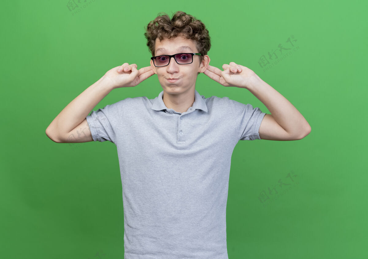 男人戴着黑色眼镜 穿着灰色马球衫的有趣的年轻人 站在绿色的墙上 露出他鼓舞人心的双耳和快乐的脸颊年轻脸颊站立