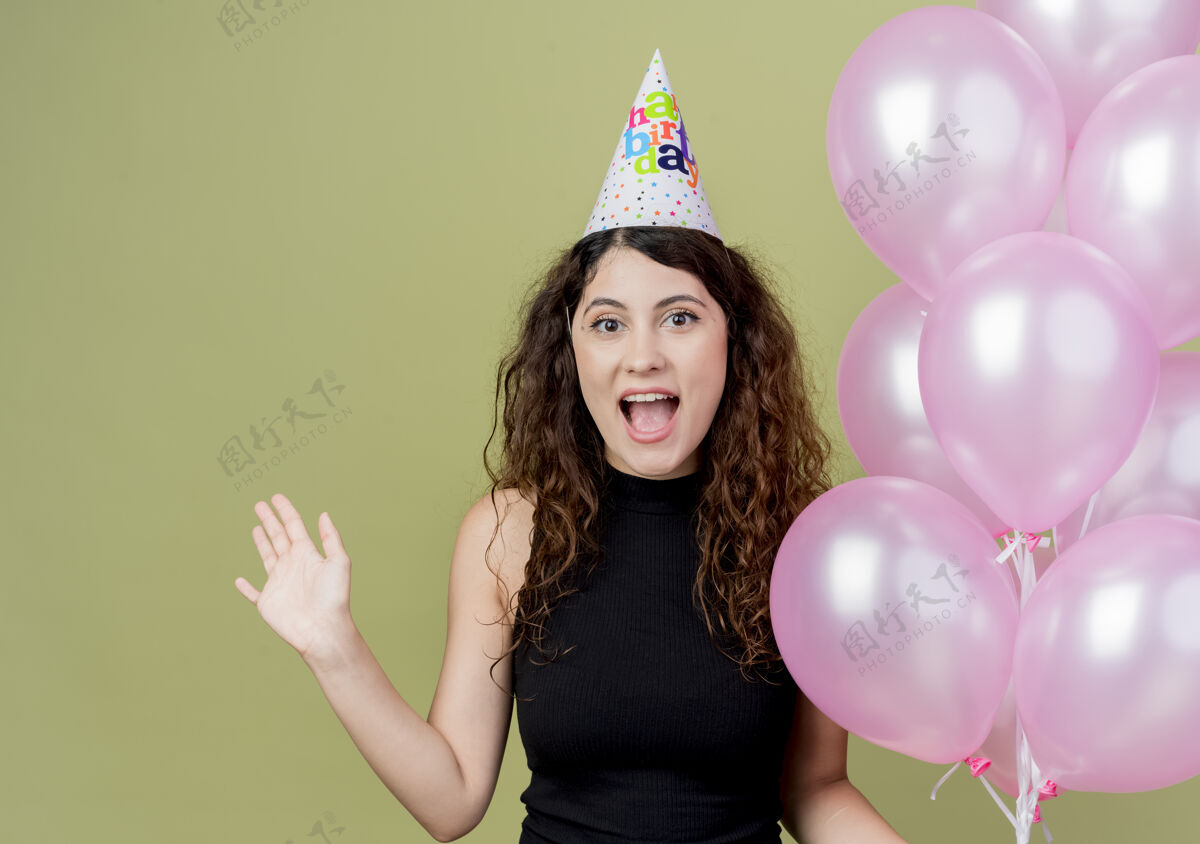 举行一位年轻漂亮的女士 卷发 戴着节日帽 手持气球 举手 高兴而兴奋地微笑着 生日派对的概念站在光墙上手空气提高