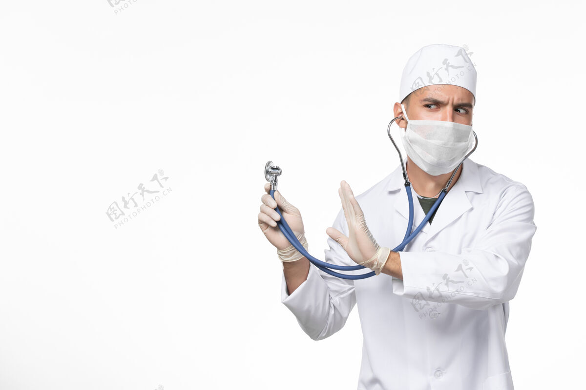 医疗正面图男医生身穿医疗服 戴口罩 因冠状病毒-使用听诊器对浅白色墙壁冠状病毒-大流行病毒疾病外套病毒冠状病毒预防
