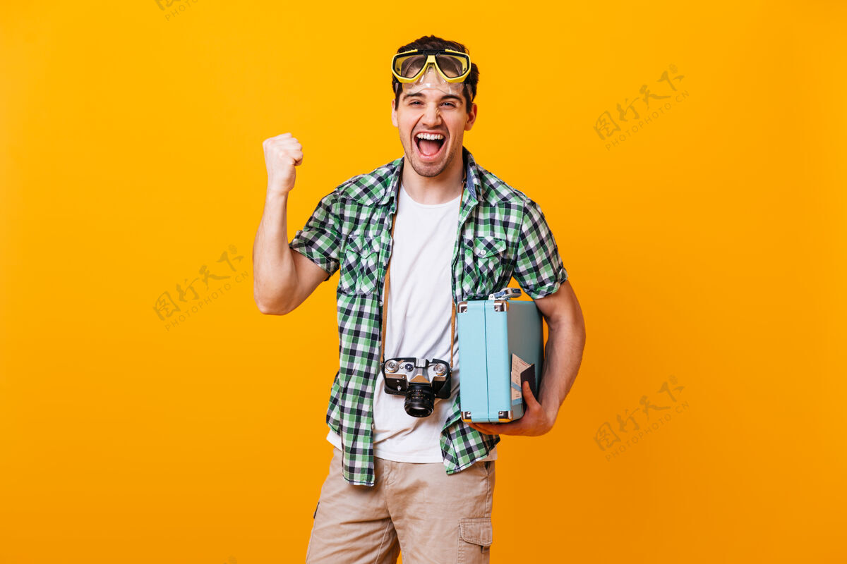 使穿着绿色衬衫和米色短裤的男人在情感上欢欣鼓舞 握紧拳头戴着潜水面具 带着复古相机和手提箱的男人在橙色的空间里欢笑机票积极采取