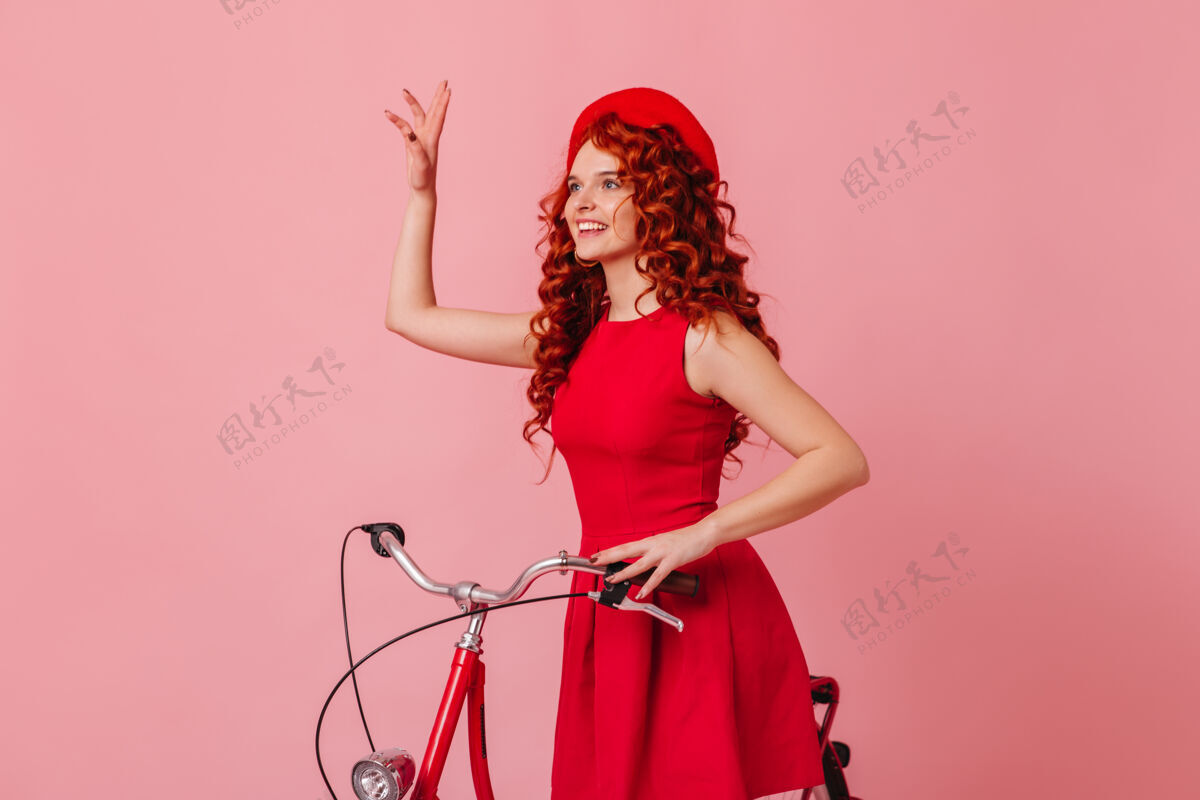 女性坐在粉色空间的自行车上挥手致意的女人戴着红色贝雷帽和裙子的女人姜黄色酷性感