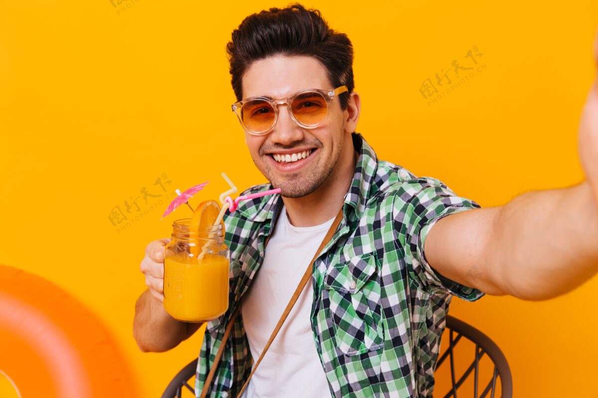 男孩年轻的黑发小伙子穿着绿色衬衫 戴着橙色眼镜 喜欢鸡尾酒 在与世隔绝的空间里自拍手机太阳镜背包