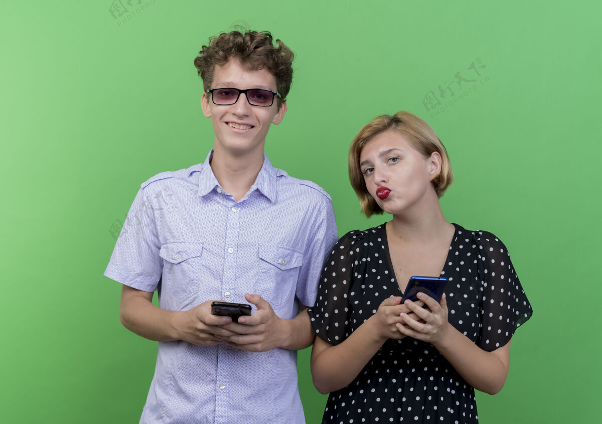移动年轻漂亮的夫妇手持智能手机微笑着站在绿色的墙上站着男人微笑