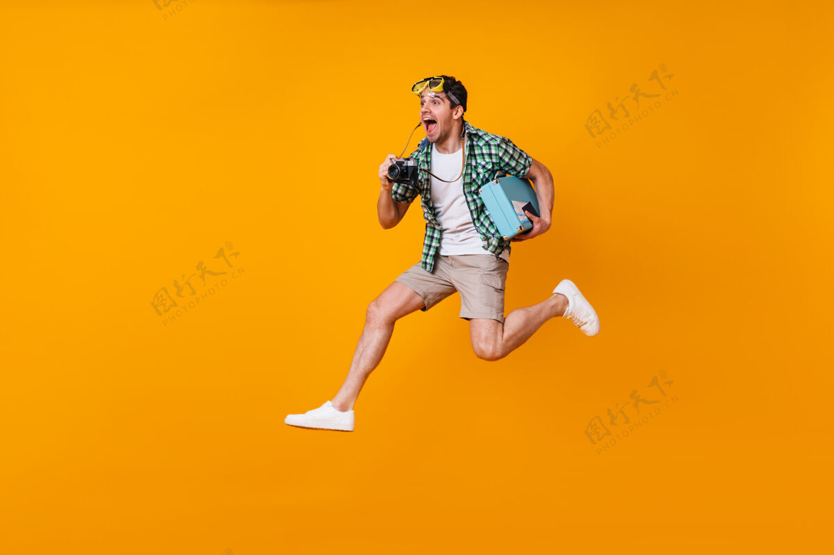 积极头上戴着潜水面具的积极男人用复古相机拍照穿着短裤和绿色衬衫的家伙拿着手提箱在橙色的空间里跳跃潜水面具使时髦