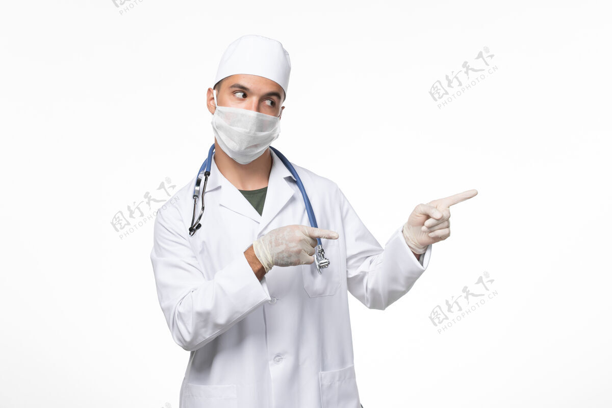 疾病正面图：男医生穿着医疗服 戴着防毒面具 白色办公桌上带着蓝色听诊器的科维德-疾病大流行病毒视图办公桌防护