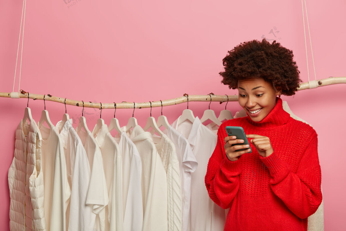 手机快乐的美国黑人网店经理 咨询客户 输入评论 销售白色的新系列 穿着红色针织毛衣 站在货架旁 隔离在粉红色的墙上购物者套装购买