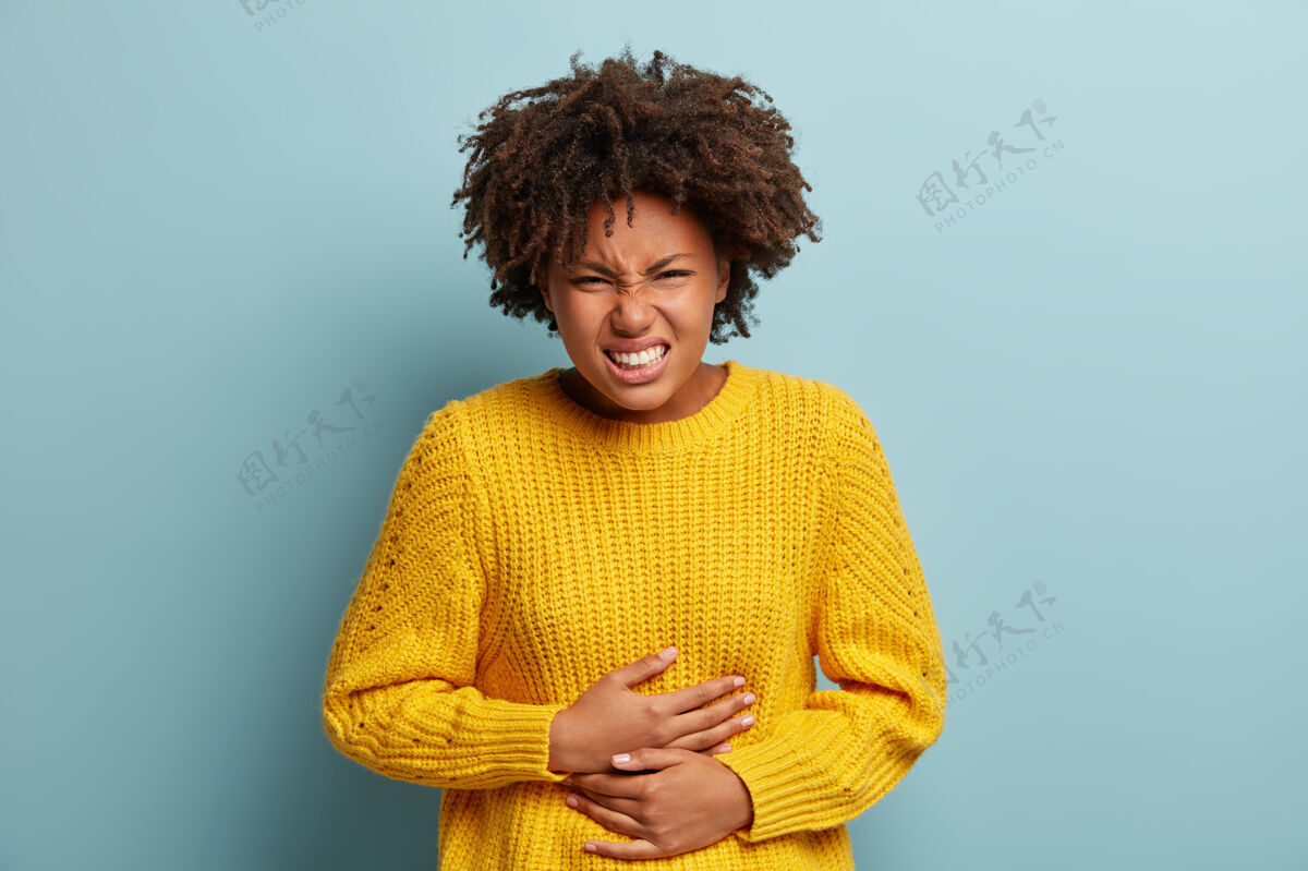 疼痛不高兴的黑皮肤女人肚子痛 经期抽筋 手放在肚子上 穿着黄色针织毛衣 笑脸 站在蓝色背景下疾病月经一