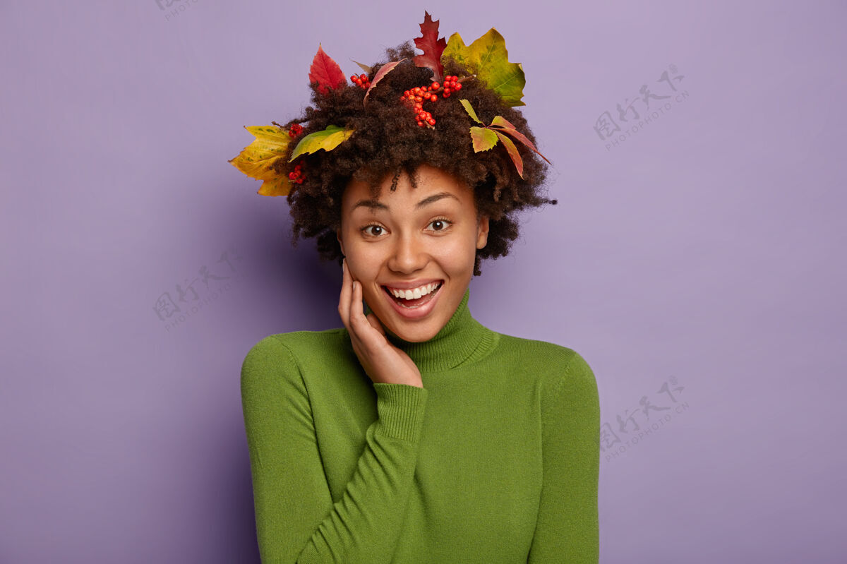 一个可爱开朗的非洲女人的肖像手放在脸颊上 穿着绿色高领毛衣 深色卷发中有叶子 笑容灿烂 露出洁白的牙齿 有着温柔的神情橙色秋天女人