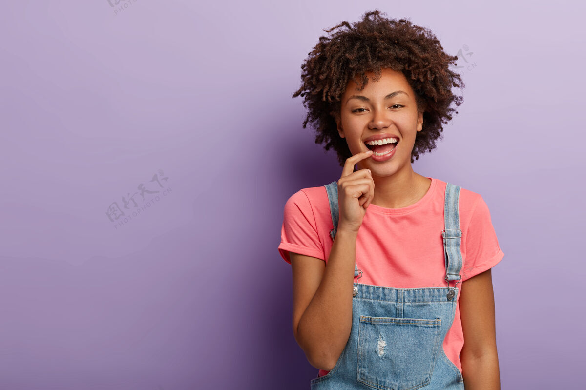美式快乐时尚的女人微笑着无忧无虑 穿着t恤和牛仔工作服 把手指放在嘴唇上 孤立在紫色的背景下T恤粉色欢呼