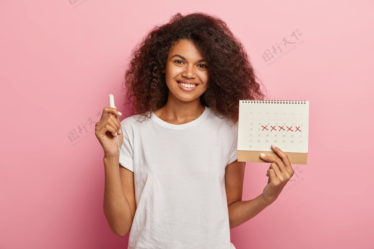 卷发快乐的卷发女士拿着月经日历 上面写着经前综合症的日子和卫生棉条 穿着休闲的白色t恤 隔离在粉色背景上日历微笑干净