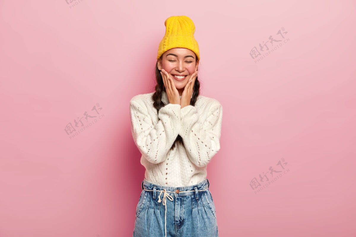 女人迷人的韩国年轻女子摸着脸颊 有着满意的表情 闭着眼睛 害羞 戴着黄色的帽子和针织毛衣套头衫一件女孩