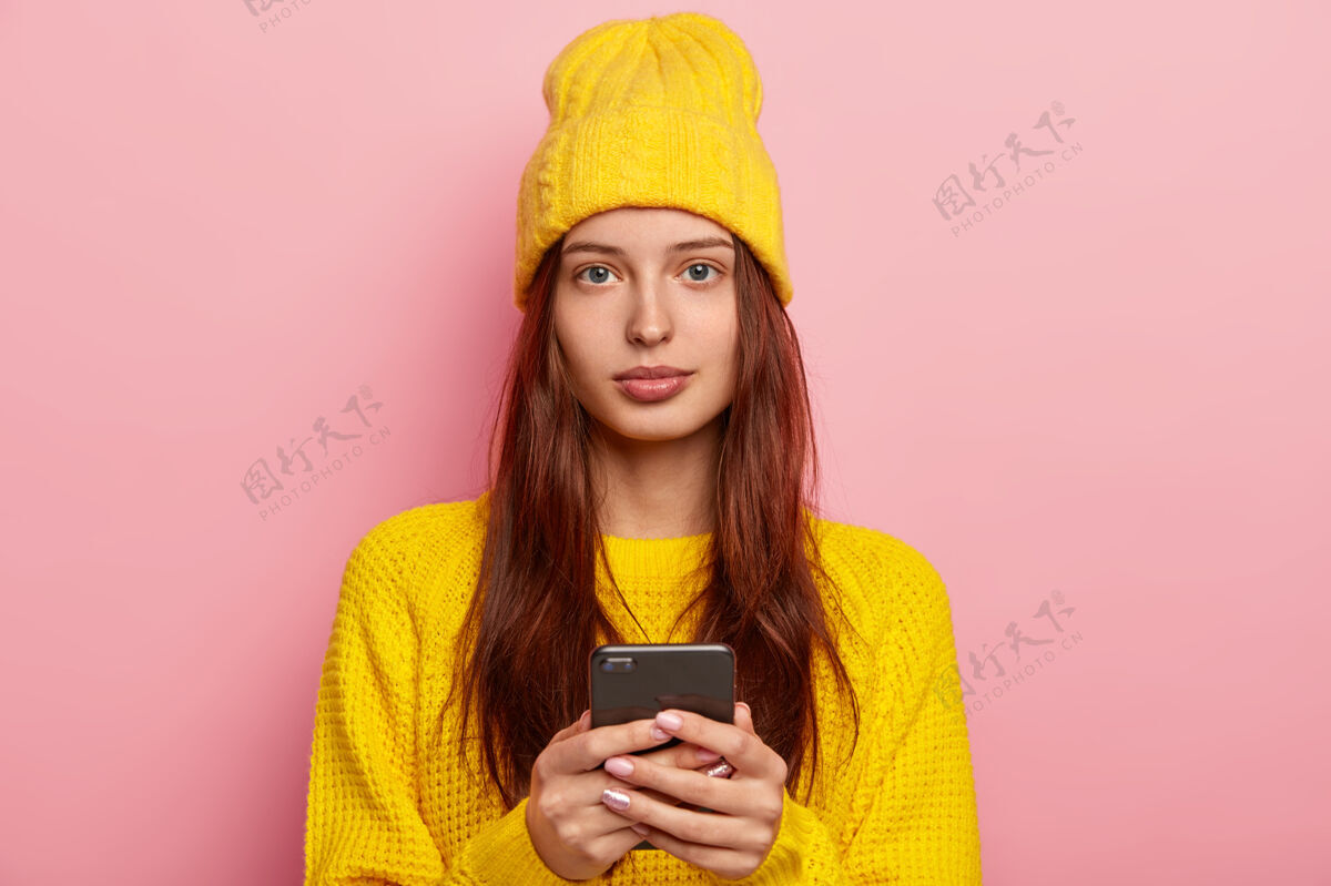 黄色容貌姣好的女人的肖像看起来很严肃 使用现代手机 戴黄色帽子和冬季毛衣 在玫瑰色背景下摆姿势 不化妆女孩表情休闲