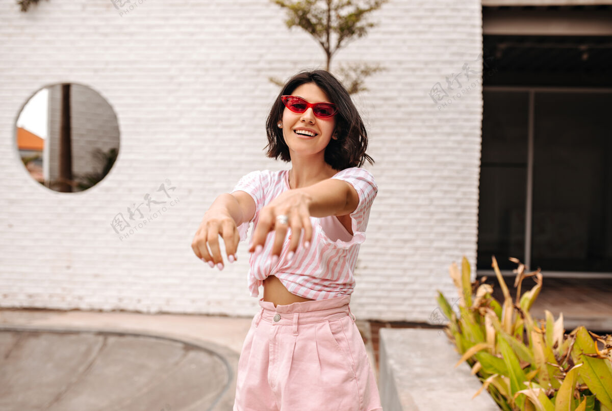 休闲戴着太阳镜的快乐女人伸出双手在街上摆姿势穿着粉色t恤的晒黑女人的户外照片户外建筑街道
