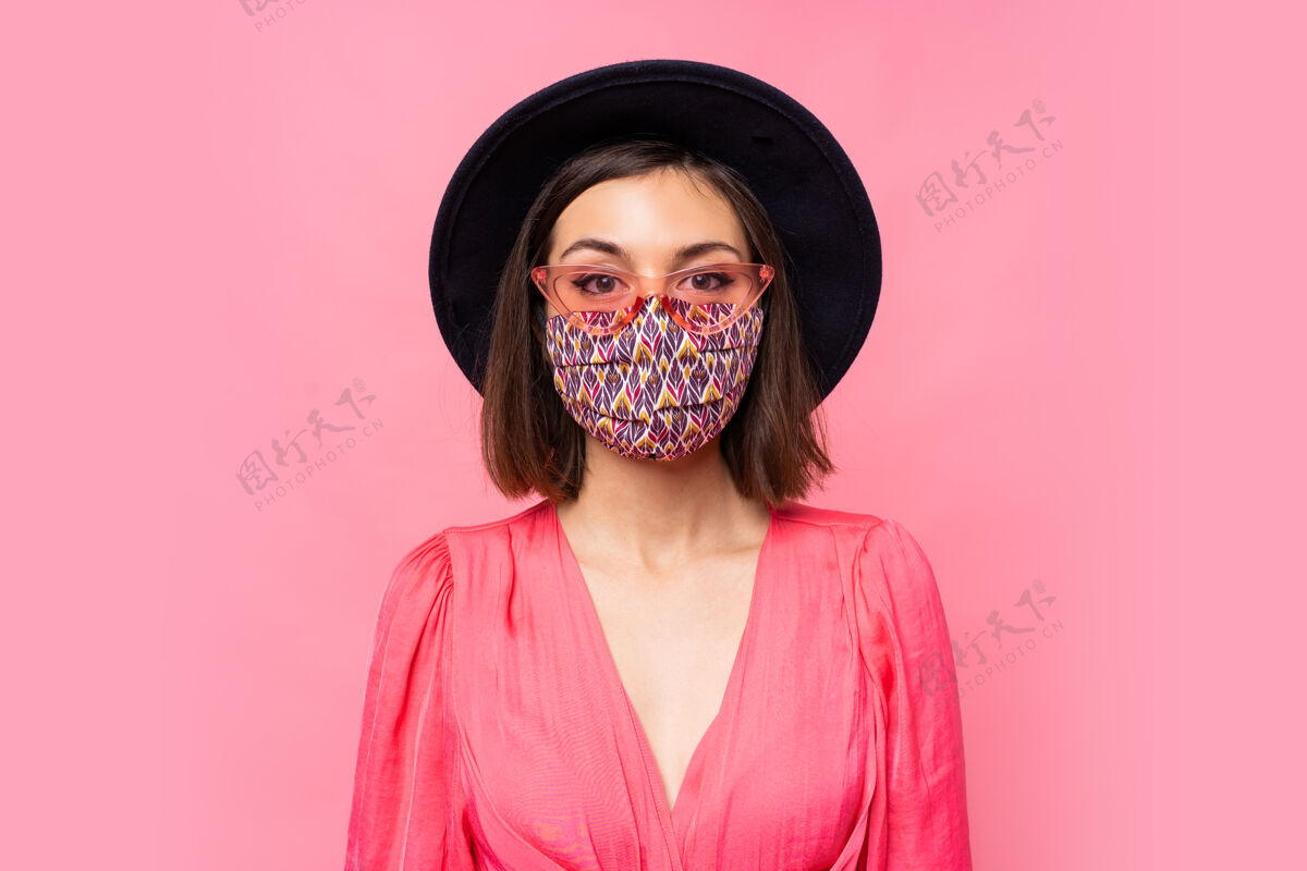 肖像欧洲模特戴着保护性的时尚面罩戴着黑色帽子和太阳镜在粉红色的墙上摆姿势女性防护距离