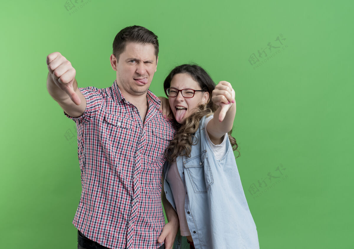站一对穿着休闲服的年轻夫妇不高兴地竖起大拇指站在绿色的墙上情侣衣服女人