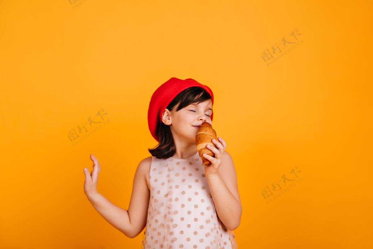 羊角面包无忧无虑地吃羊角面包的女婴可爱的孩子站在黄色的墙上零食帽子休闲