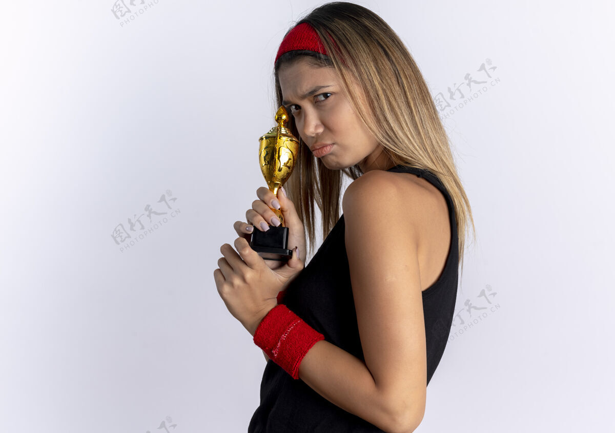 年轻愤怒的年轻健身女孩身穿黑色运动服 头戴红色头巾 手持奖杯站在白墙上怨恨看头带