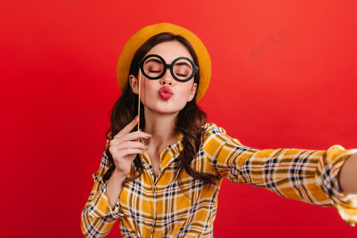 喜悦戴着帽子的快乐女孩把眼镜放在棍子上送吻穿黄衬衫的年轻人在红墙上自拍魅力可爱模型