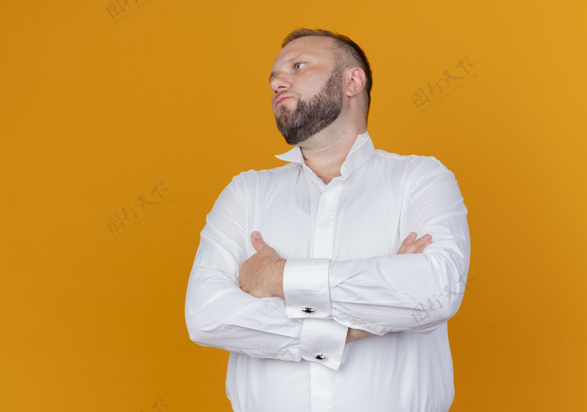 靠边一个留着胡子的男人穿着白衬衫 双手交叉放在胸前 站在橙色的墙上 向旁边看男人胸十字架