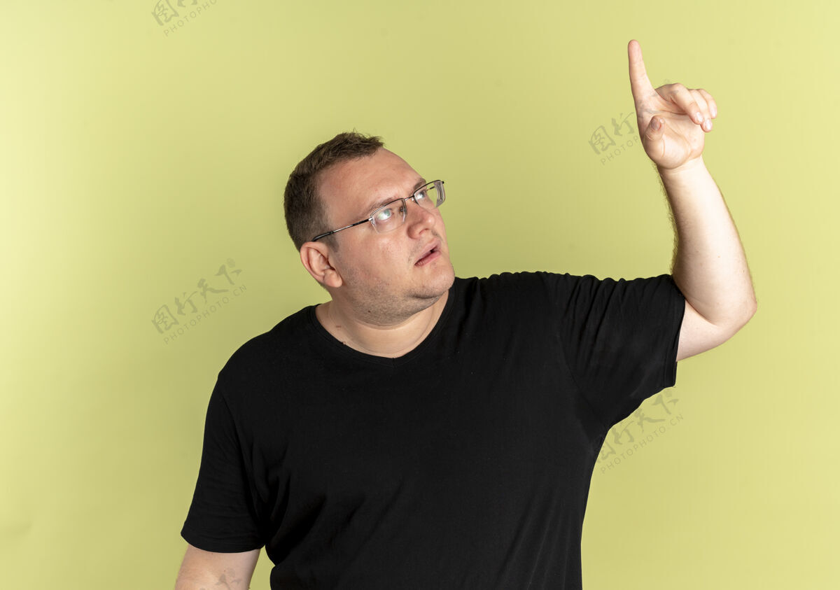 东西一个戴眼镜 身穿黑色t恤的超重男子抬起头 严肃地用食指指着某个东西 站在浅色的墙上光指向手指