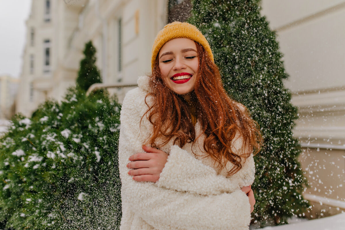 脸快乐的姜黄色女士在雪下摆姿势微笑可爱的女士戴着针织帽子站在云杉旁边服装户外成人