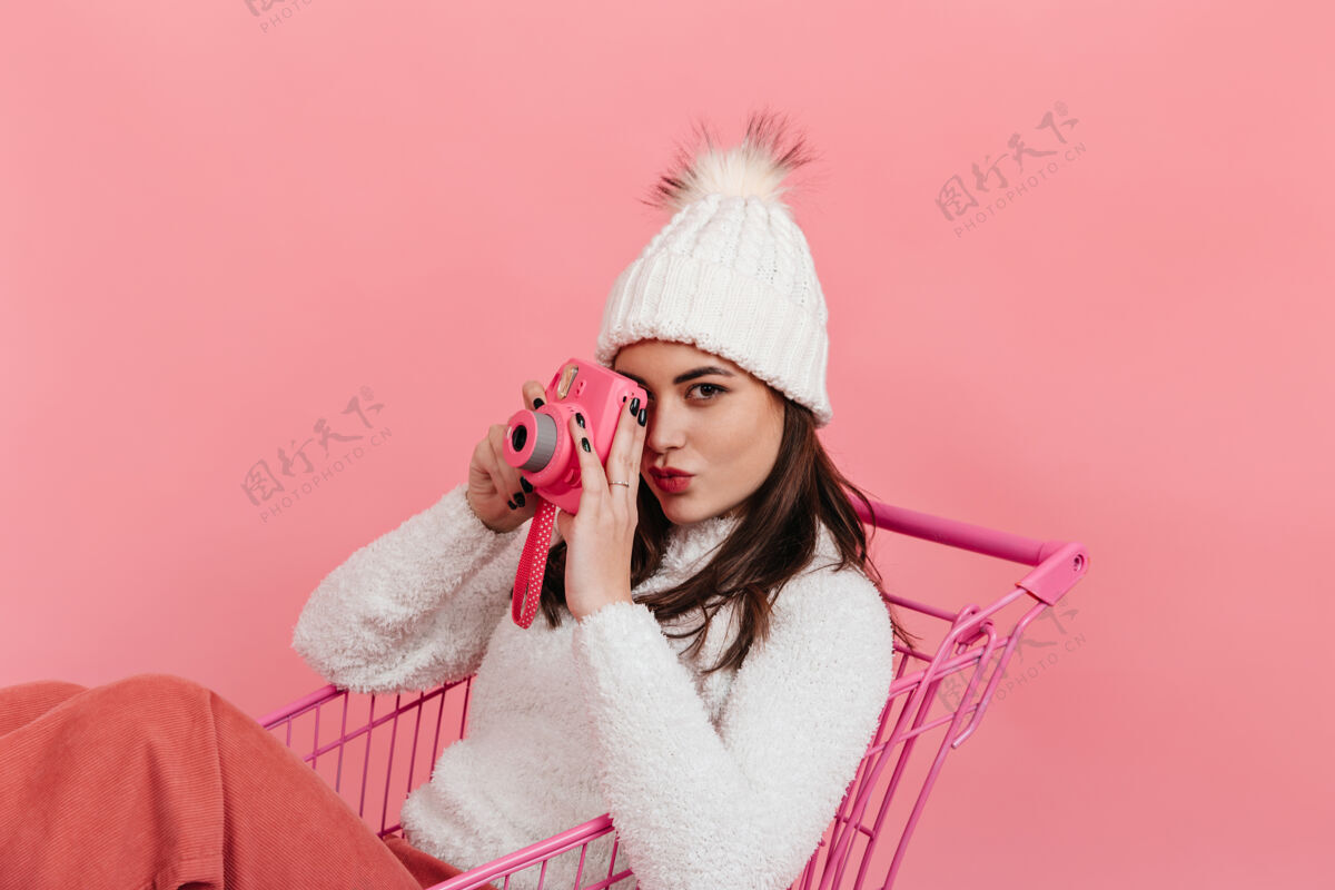人一张棕色眼睛 戴着白色帽子 穿着毛衣的女士坐在粉色墙上的超市手推车里 在instax上拍照的照片欢呼白色毛衣学生