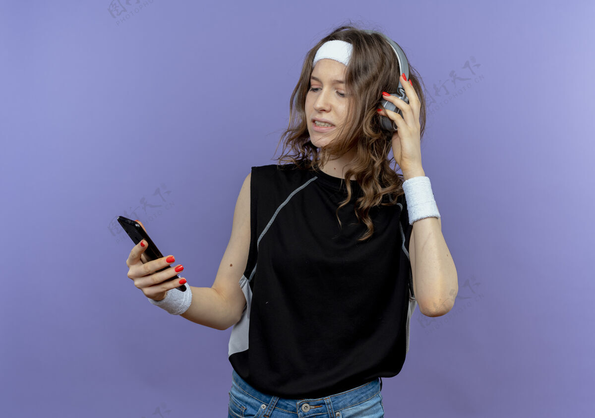 年轻身穿黑色运动服的年轻健身女孩戴着头带和耳机 站在蓝色的墙上看着她的智能手机搜索音乐音乐耳机女孩