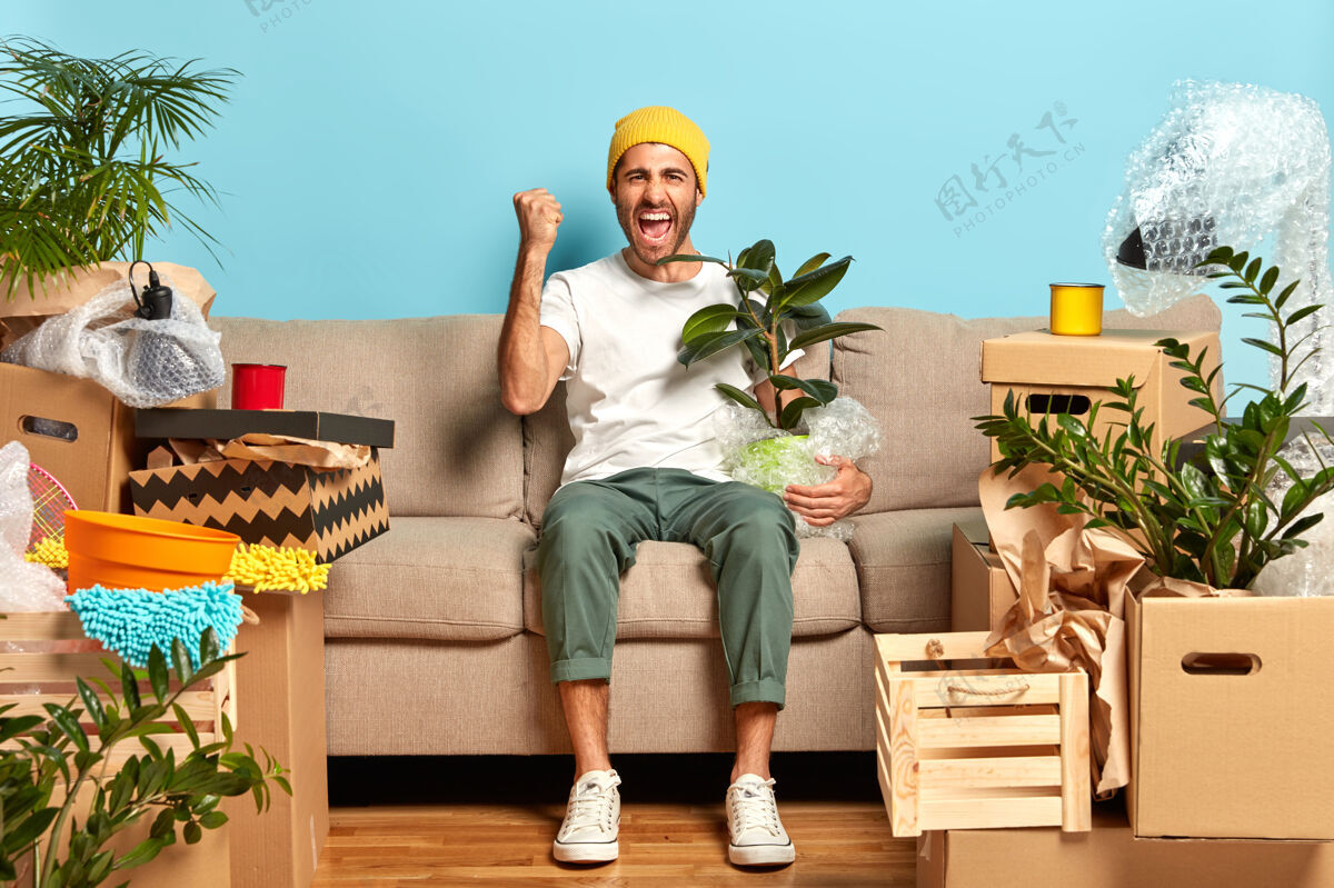 舒适喜出望外的家伙握紧拳头 在沙发前摆姿势 抱着包好的盆栽放松帽子坐着