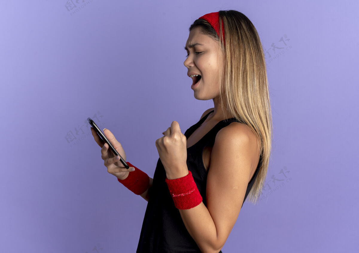 屏幕身穿黑色运动服 头戴红色头巾的年轻健身女孩看着自己的智能手机屏幕 握紧拳头站在蓝色的墙上 快乐而兴奋拳头女孩握紧