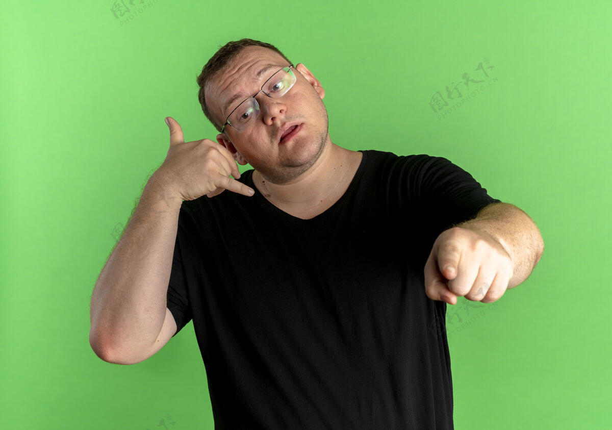 站立戴眼镜的超重男子身穿黑色t恤 用食指指着绿色的墙壁做着“叫我”的手势指点手势制造