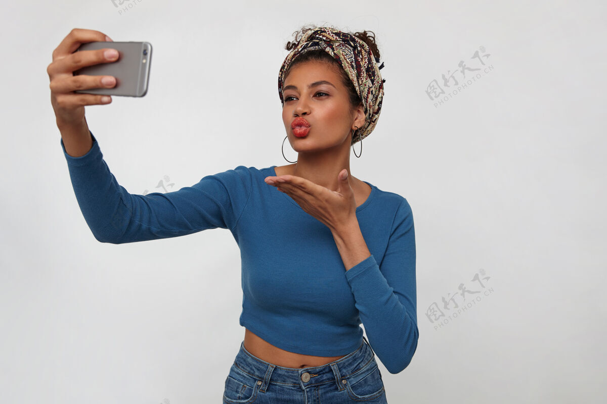 深色迷人的年轻黑发黑皮肤女性 站在白色背景上 一边用手机拍摄自己的照片 一边对着镜头送上飞吻戒指长移动