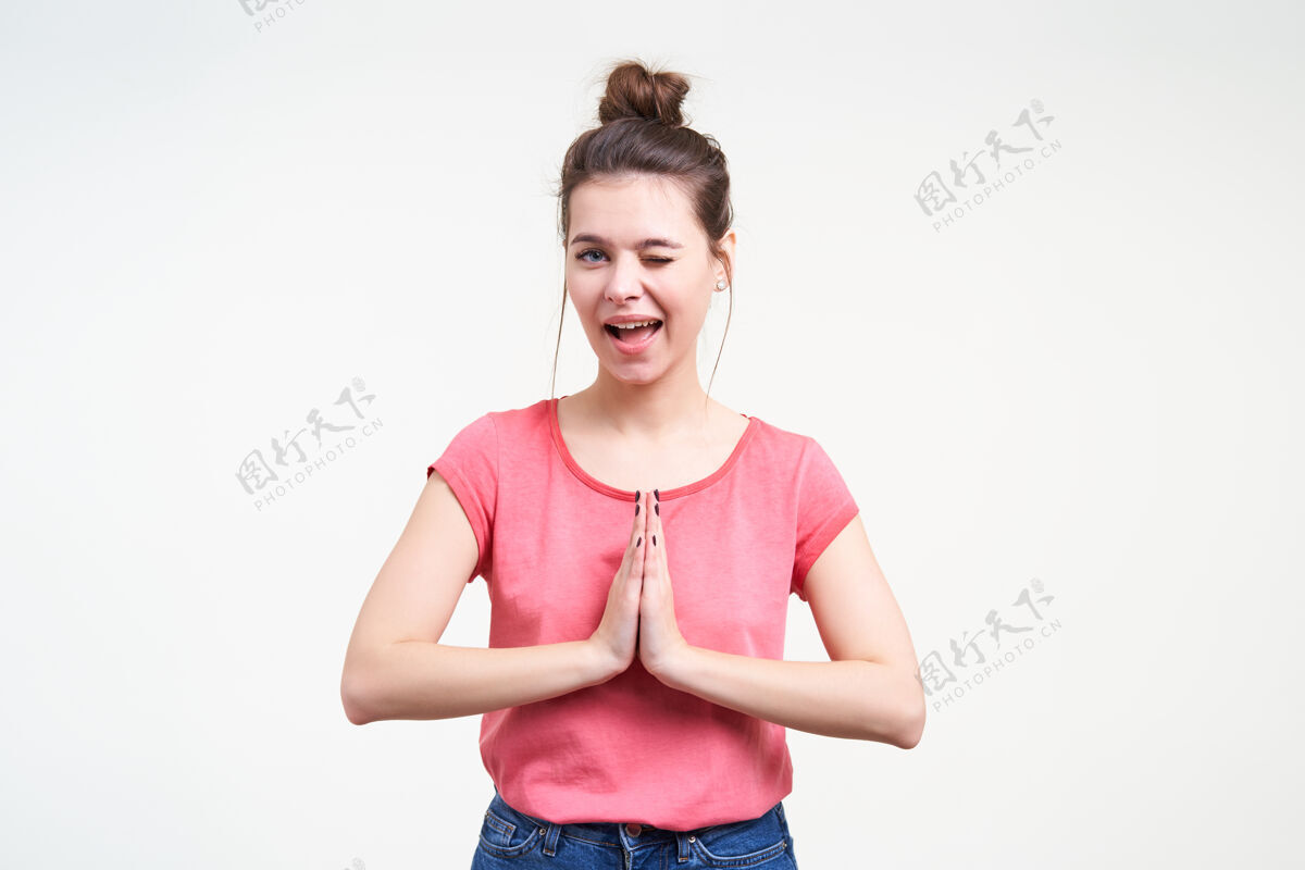 结快乐的年轻可爱的黑发女性闭上一只眼 顽皮地看着相机 双手合十做着祈祷的手势 孤立在白色的背景下积极女士表情